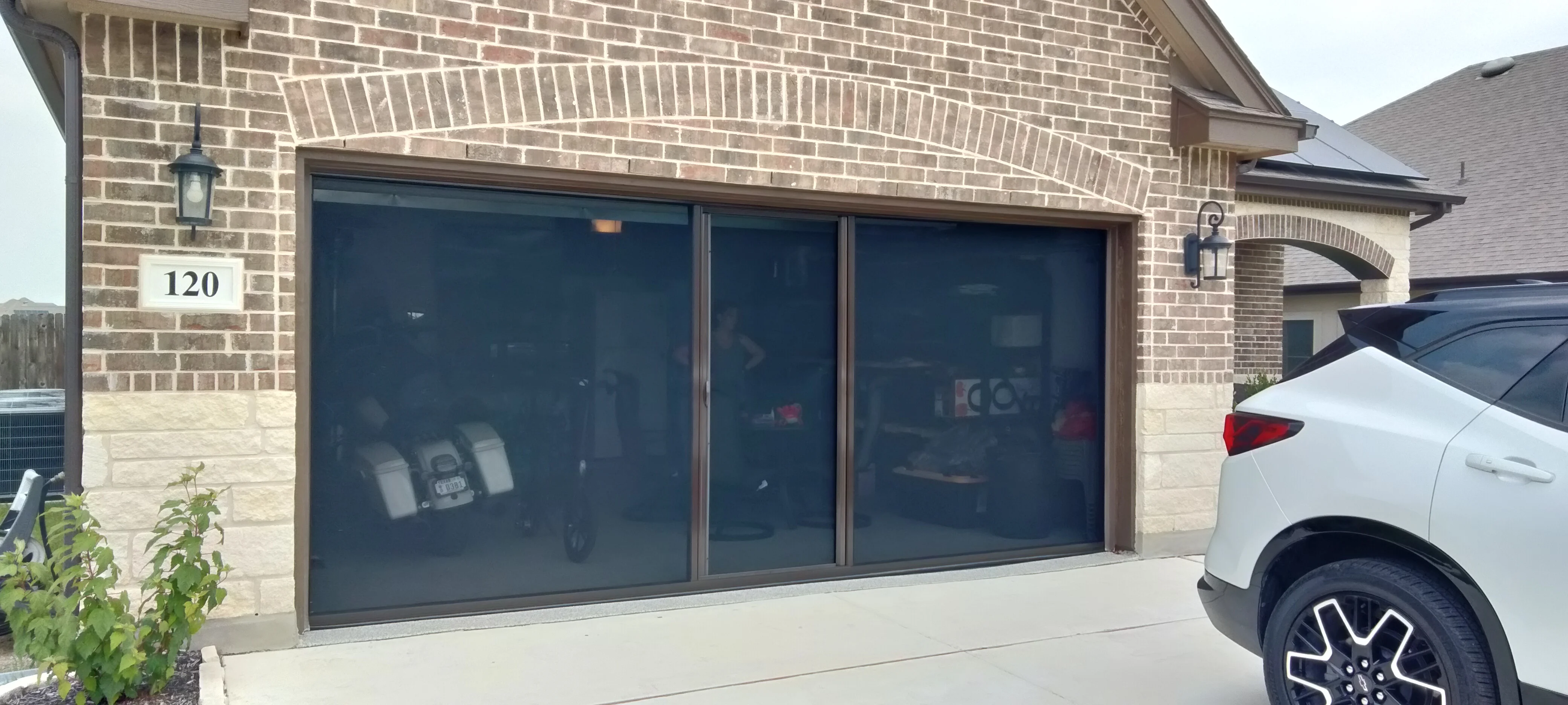 Three-panel dark-tinted garage screen door with bronze frame on brick home exterior